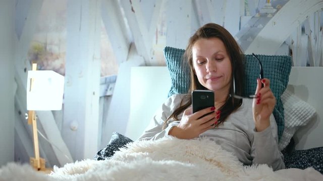 Woman in glasses lies in a bed and uses a smartphone before bedtime
