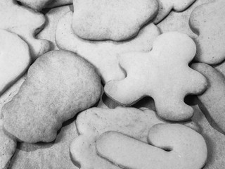 A close up black and white food photograph of a pile of baked sugar Christmas cookies in various shapes including gingerbread man, candy canes and snowman on a metal baking pan or sheet.