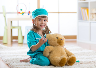 Happy child little doctor girl examines teddy bear in nursery room at home.