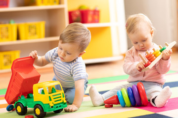 Kids playing with educational toys. Children sit on a rug in a play room at home or kindergarten. Toddler boy with toy lorry and baby girl with rings.