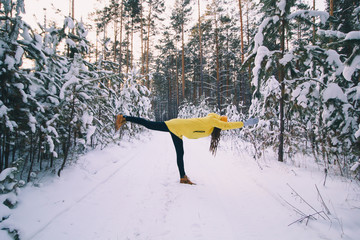 beautiful young woman in yellow jacket and hat doing workout or yoga in winter park among snow covered trees, healthy lifestyle