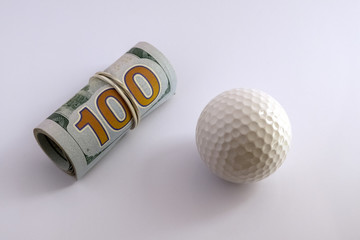Golf ball next to a roll of hundred dollar bills of the USA with a white rubber band. The concept of sports betting, golfing competitions for money or corruption.