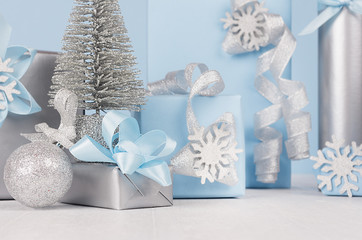 Elegance soft christmas home decorations - silver small fir with blue and metallic gift boxes with shiny ribbons and snowflakes closeup on light white wood board and pastel blue wall.