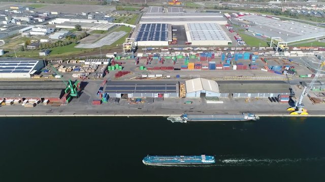 Aerial footage of harbor area industrial zone small barge moving west over canal in background showing the port buildings and docked ship loading freight for transportation 4k high resolution