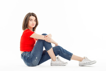 Side view of casual teenage brunette in denim and red t-shirt sitting isolated on white background looking at camera