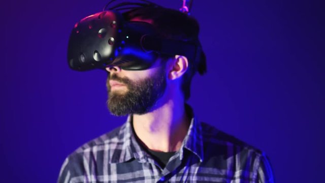 Close-up portrait of bearded attractive man wearing vr glasses looking around at the Metaverse, blue background, virtual reality concept