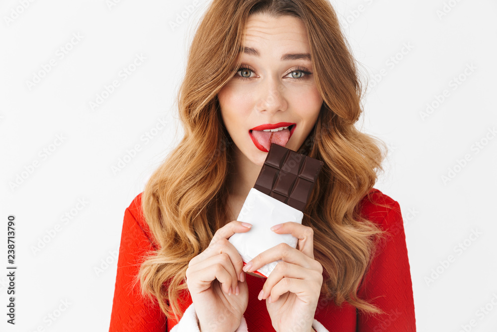 Poster Portrait of caucasian woman 20s wearing Santa Claus red costume smiling and eating chocolate bar, isolated over white background