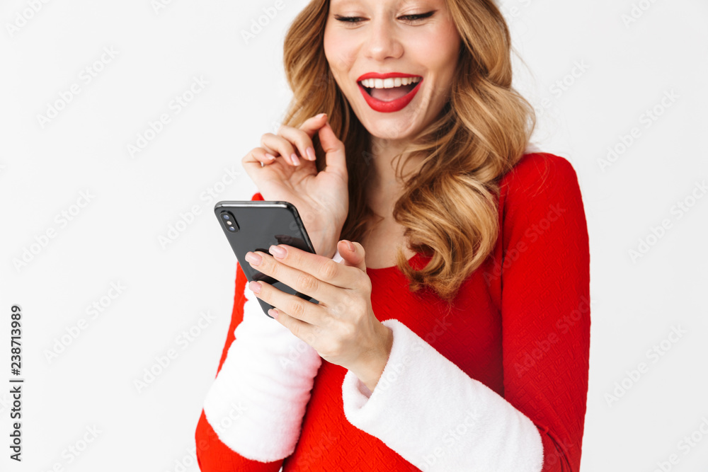 Sticker Portrait of happy woman 20s wearing Santa Claus red costume smiling and holding black smartphone, isolated over white background