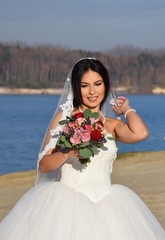 Fototapeta na wymiar Wunderschöne Braut in einem weißen Brautkleid, Schleier, und Blumenstrauß an einem Strand bei blauem Himmel im Sommer