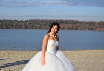 Fototapeta na wymiar Wunderschöne Braut in einem weißen Brautkleid, Schleier, und Blumenstrauß an einem Strand bei blauem Himmel im Sommer