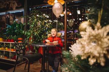 Happy and curious child sitting with air baloon in hand. Warmly dressed. Christmas decorations and home plants around him. Beautifully decorated christmas tree and toy santa.