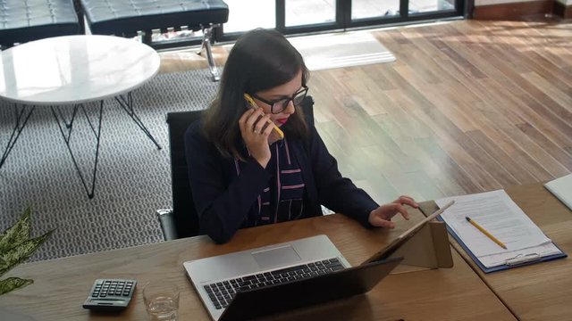 High angle view of Caucasian business lady sitting at working table, talking on phone, swiping some information on tablet, then putting away smartphone and starting typing on laptop&rsquo;s keyboard