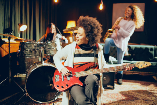 Girls playing jazz music. In foreground one woman playing bass guitar and in background other two playing saxophone and drums. Home studio interior.