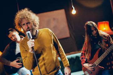 for the gig. Male singer with curly hair holding microphone and singing. In background band playing instruments. Home studio interior.