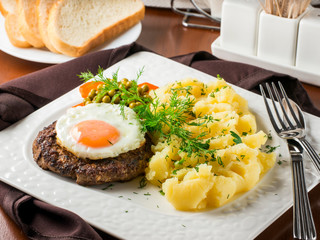 Beefsteak with fried egg and mashed potatoes on white plate close-up on dark brown wooden table in restaurant