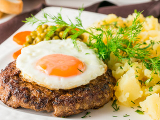 Beefsteak with fried egg and mashed potatoes on white plate close-up on dark brown wooden table in restaurant