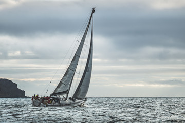Fototapeta premium Close-up lone yacht sailing in the sea. Northern Ireland. Blue grey cloudy sky background. Outdoor tourist activity. Wild nature environment before the rain. Mysterious Irish ocean scape.