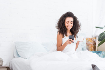 Obraz na płótnie Canvas smiling curly girl with coffee cup using smartphone in bed during morning time at home