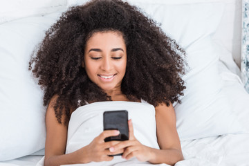 happy african american young woman using smartphone in bed during morning time at home