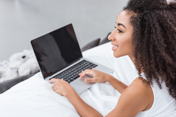 side view of african american curly girl using laptop with blank screen in bed during morning time at home