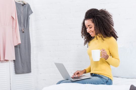 joyful african american female freelancer with coffee cup working on laptop at home