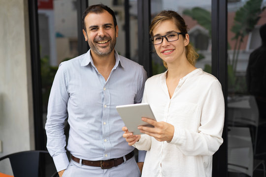 Portrait of happy mid adult businessman and young businesswoman wearing glasses standing together, looking at camera and smiling. Woman holding digital tablet. Team concept