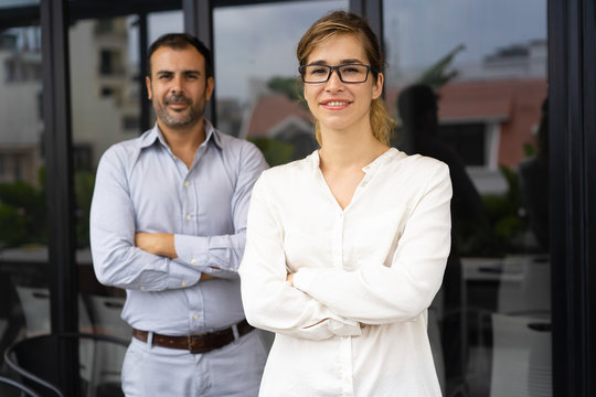 Portrait of smiling successful female leader wearing glasses, her mid adult male colleague standing behind. Businesspeople posing in office. Female leader concept