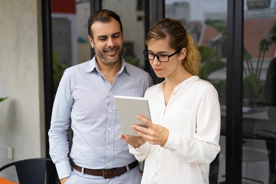 Serious female trainee showing her work to executive on digital tablet. Mid adult Caucasian businessman looking at pc and smiling. Apprentice concept