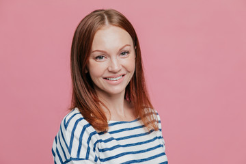 Fototapeta premium Sideways shot of pleasant looking happy brown haired woman with straight hair, healthy skin, pleasant smile, dressed in casual striped sweater, isolated over pink background. Emotions concept