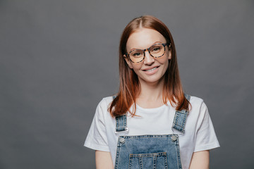 Isolated shot of positive pretty woman tilts head, has charming smile, wears glasses, white t shirt and overalls, poses over grey background with free space for your advertising content or promotion