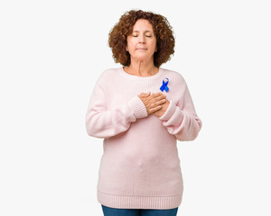 Middle ager senior woman wearing changeable blue color ribbon awareness over isolated background smiling with hands on chest with closed eyes and grateful gesture on face. Health concept.