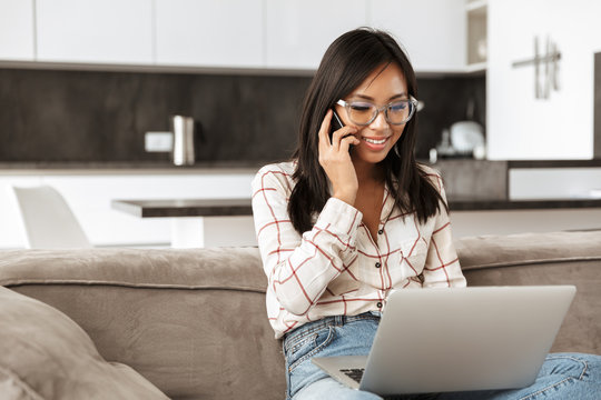 Photo Of Cute Asian Woman 20s Speaking On Cell Phone And Working On Laptop, While Sitting At Sofa In Apartment