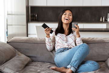 Photo of happy asian woman 20s holding remote control and cup, while sitting at sofa in cozy apartment