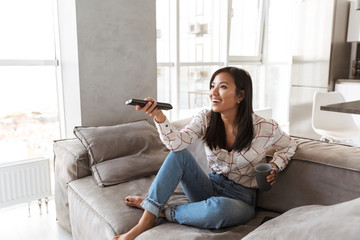 Photo of fashion asian woman 20s holding remote control and watching TV, while sitting at sofa in...