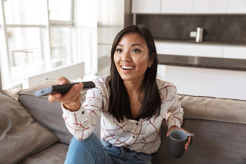 Photo of positive asian woman 20s holding remote control and watching TV, while sitting at sofa in cozy apartment