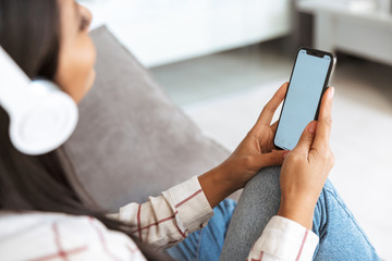 Photo of young asian woman 20s wearing headphones holding smartphone, while sitting on sofa in living room