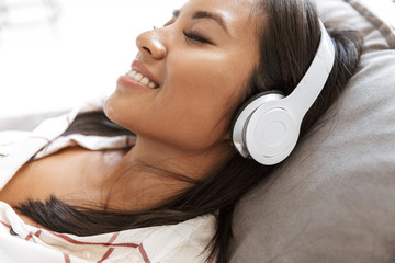 Photo of joyful asian woman 20s wearing headphones listening to music, while lying on sofa in living room