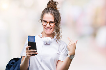 Young brunette student girl wearing backpack, headphones and smartphone over isolated background with surprise face pointing finger to himself