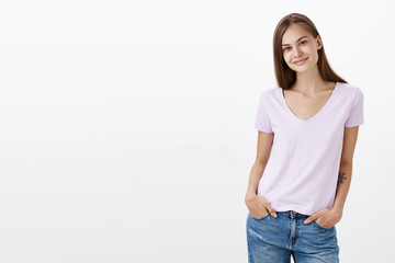 Fototapeta premium Charming shy and cute european young female brunette in casual blouse standing with hands in pockets over white background tilting head and smiling with friendly look getting know new coworkers