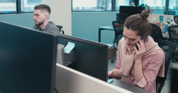 Happy young smiling Caucasian woman sitting at her desk in the office working and answering a phone call. 4K UHD