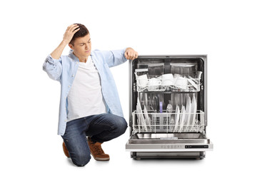 Angry young guy kneeling next to a loaded dishwasher holding his head