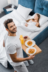 overhead view of smiling man holding wooden tray with breakfast while woman sleeping in bed