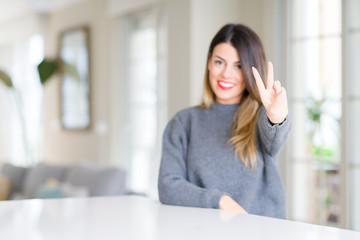 Obraz premium Young beautiful woman wearing winter sweater at home smiling looking to the camera showing fingers doing victory sign. Number two.
