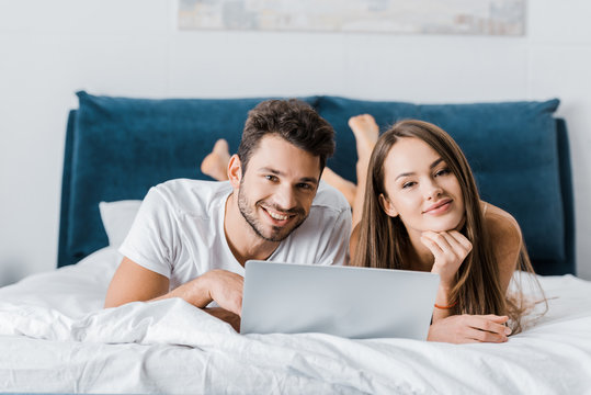 Young Smiling Couple Lying In Bed With Laptop And Looking At Camera