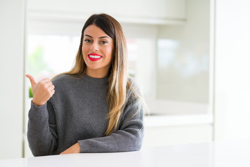 Young beautiful woman wearing winter sweater at home smiling with happy face looking and pointing to the side with thumb up.