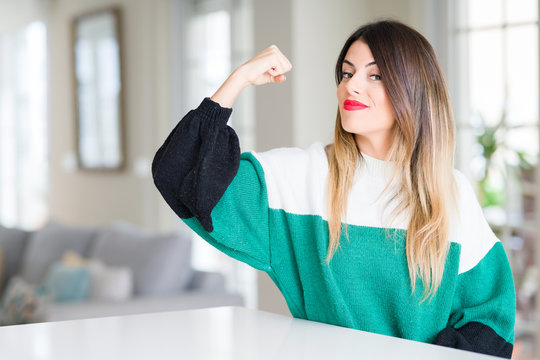 Young beautiful woman wearing winter sweater at home Strong person showing arm muscle, confident and proud of power