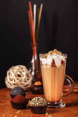 Beautifully designed latte macchiato coffee with whipped cream and sprinkled with crushed halva, in high transparent glass, on a wooden table, on a dark background with cakes