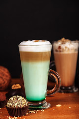 Coffee latte macchiato with mint syrup and whipped cream, in high transparent glasses, on a wooden table, on a dark background with cakes