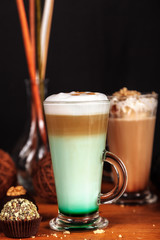 Coffee latte macchiato with mint syrup and whipped cream, in high transparent glasses, on a wooden table, on a dark background with cakes