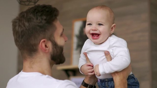 Father plays with infant son in white t-shirt looks at him and laughs. Laughing baby looking at the camera. Loving father